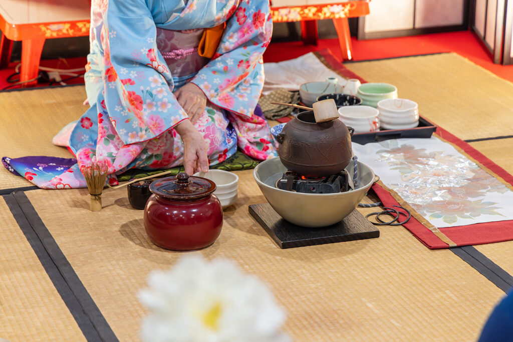 Japanese tea ceremony demonstrated by a master wearing a traditional kimono at an Asian exhibition with tea utensils, an iron kettle on a stove, and ceramic bowls on a tatami mat